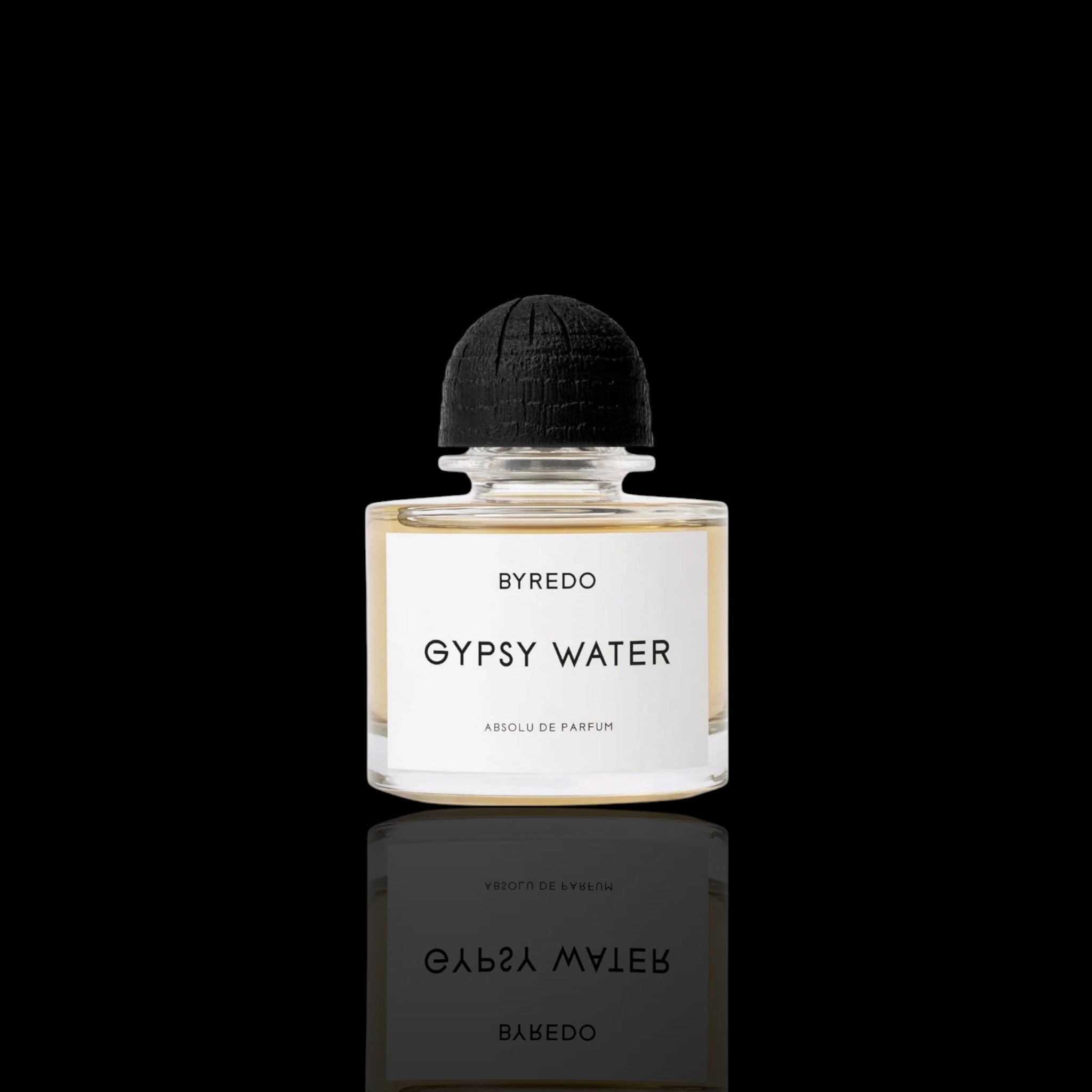 A bottle of Byredo Gypsy Water Absolu de Parfum centered on a solid black background with a clear reflection below. The bottle is clear glass containing a pale golden liquid, featuring a white rectangular label with black text and a textured black dome cap.