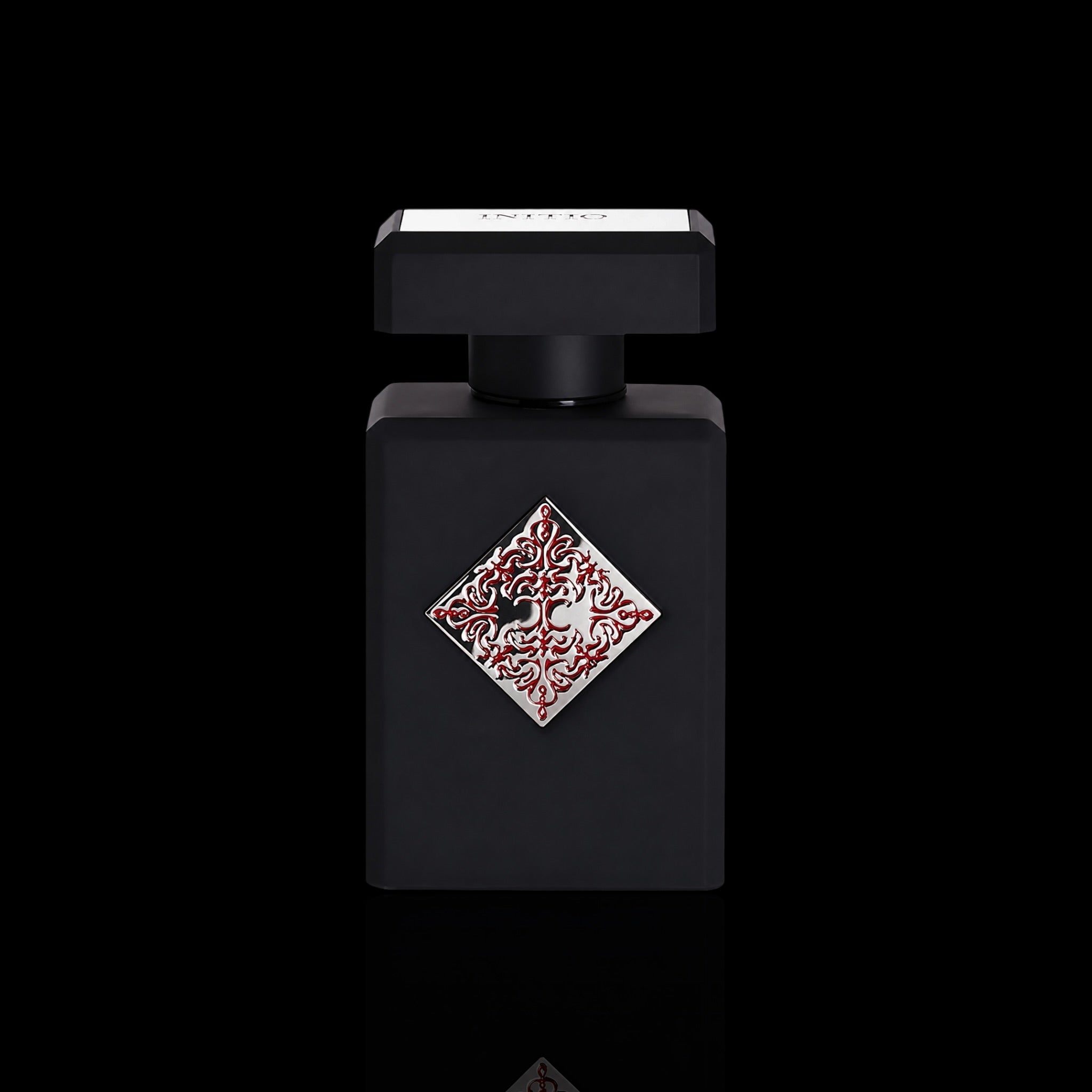 A matte black rectangular Initio absolute aphrodisiac perfume bottle with a square cap, featuring a decorative diamond-shaped silver and red patterned emblem on the front, set against a solid black background.